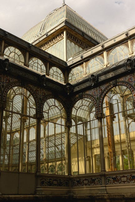 A detailed exterior view of a historic glass and wrought iron conservatory, showcasing multiple large arched windows with gold-colored frames and decorative ironwork. The structure features ornate met