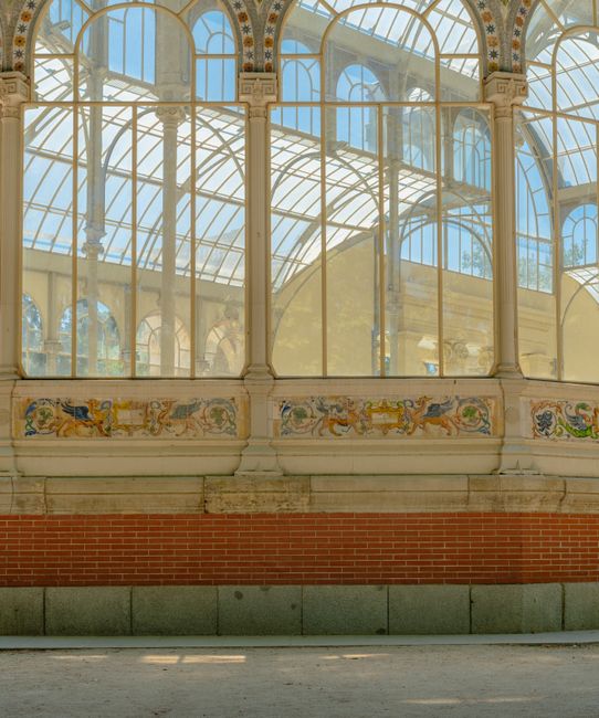 A detailed view of an ornate glasshouse with large, arched windows and decorative painted panels beneath the glass structure. The glass panes are supported by white metal frameworks, allowing ample natural light to illuminate the interior. The lower part of the structure features a brick base with a row of stone panels above it. Inside, there are wooden furniture pieces, cardboard boxes, and plastic wrapping materials arranged in a spacious area, suggesting an ongoing home relocation or furniture transport process. A moving trolley and packing blankets are visible, indicating a professional move by Crystal Palace Man and Van, as part of their house removals and packing and moving services. The scene captures both the architectural elegance of the building and the logistical activity involved in home relocation within a historic or conservatory-style environment.