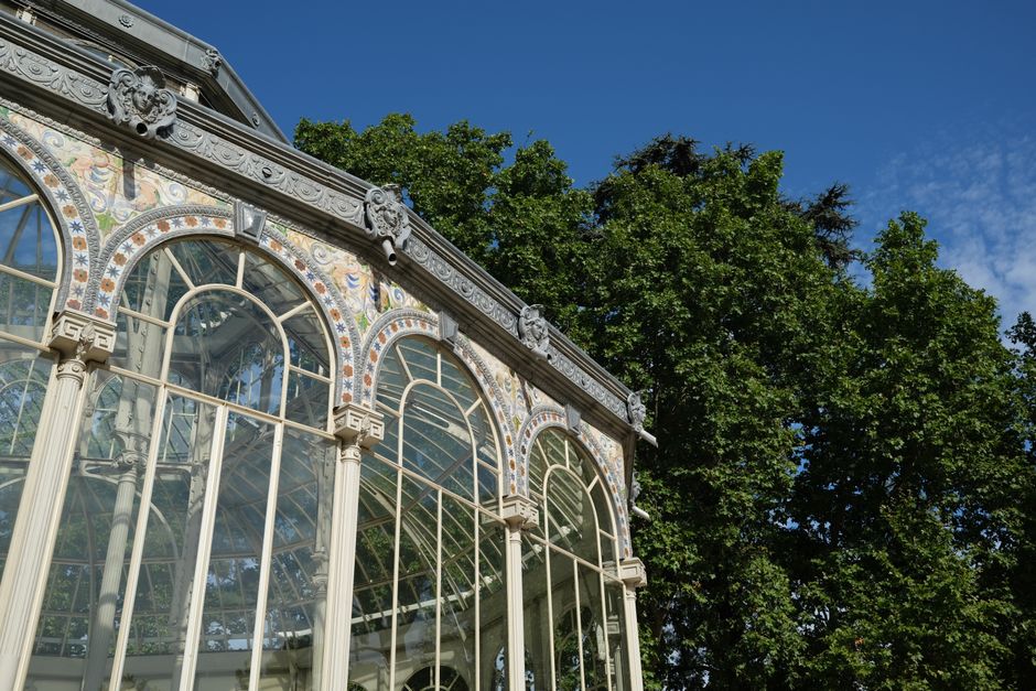 A detailed view of an ornate glasshouse with large, arched windows and decorative painted panels beneath the glass structure. The glass panes are supported by white metal frameworks, allowing ample natural light to illuminate the interior. The lower part of the structure features a brick base with a row of stone panels above it. Inside, there are wooden furniture pieces, cardboard boxes, and plastic wrapping materials arranged in a spacious area, suggesting an ongoing home relocation or furniture transport process. A moving trolley and packing blankets are visible, indicating a professional move by Crystal Palace Man and Van, as part of their house removals and packing and moving services. The scene captures both the architectural elegance of the building and the logistical activity involved in home relocation within a historic or conservatory-style environment.