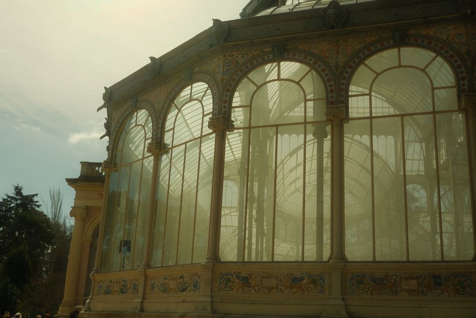 A glass conservatory with ornate yellow and gold decorative features, large arched windows, and intricate painted detailing along the lower sections. The structure is partially illuminated by sunlight