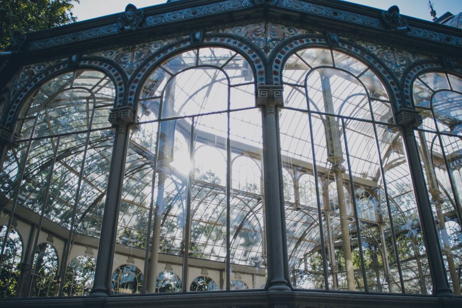 The image shows a close-up view of a historic glasshouse with ornate metal framing and large, curved glass panels reflecting sunlight, set against a bright blue sky. The structure features decorative arches and detailed metalwork along the edges of the glass panels. A tall, spire-like finial is visible atop the central dome of the conservatory. Inside the glasshouse, glimpses of plants can be seen through the transparent walls. This architectural style is typical of Victorian-era glass and iron greenhouses, associated with London's famous historic conservatories such as the Crystal Palace. The scene captures the intricate design and construction details of this classical horticultural structure, relevant for content related to house and garden preservation or restoration, as well as professional removals involving valuable or delicate architectural elements.