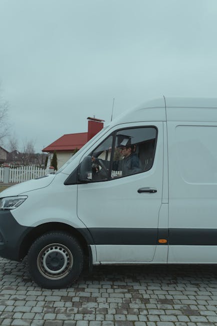 A white commercial van parked on a cobblestone driveway in a residential area, with a man seated inside the driver's seat. The van features black side mirrors, a black front bumper, and visible wheel rims. Behind the vehicle, there is a house with a red roof, a chimney, and a white picket fence along the boundary of the property. Overcast sky clouds are visible in the background, indicating a cloudy day. The scene suggests the van is used for home relocation or furniture transport, consistent with services provided by Crystal Palace Man and Van, as part of their house removals and packing and moving processes.