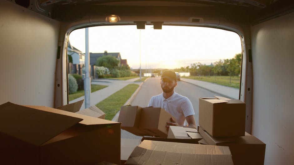 A man wearing a white polo shirt and sunglasses is seen standing outside a chalkboard driveway during golden hour, unloading or loading cardboard boxes into the back of a van, which is parked on a residential street. The open rear doors reveal several packed cardboard cartons positioned inside the van, with some placed on the vehicle's floor and others being handled by the man. The background features suburban houses, greenery, and a setting sun casting warm light across the scene. This image depicts furniture transport or packing and moving activities associated with home relocation services offered by [COMPANY_NAME], highlighting the logistical process of house removals and the careful handling of belongings during a domestic move.